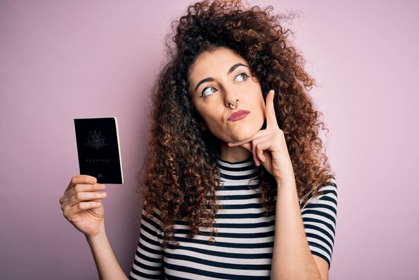 Mooie toeristische vrouw met krullend haar en piercing houden van Australië Australische paspoort id ernstige gezicht denken over de vraag, zeer verward idee - Foto, afbeelding