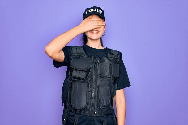 Joven mujer policía vistiendo uniforme chaleco de seguridad a prueba de balas sobre fondo púrpura sonriendo y riendo con la mano en la cara cubriendo los ojos para sorpresa. Concepto ciego
. - Foto, imagen