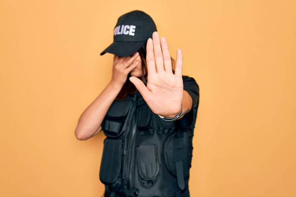 Fiatal gyönyörű barna rendőrnő, rendőri egyenruhát visel golyóálló és sapka fedi a szemét a kezét, és ezzel stop gesztus szomorú és félelem kifejezés. Zavarodott és negatív koncepció. - Fotó, kép