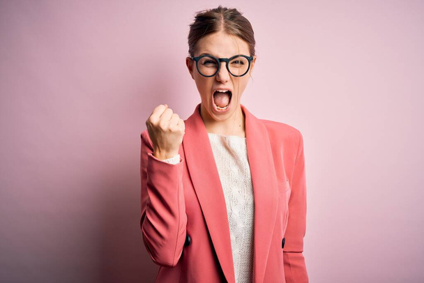 Young beautiful redhead woman wearing jacket and glasses over isolated pink background angry and mad raising fist frustrated and furious while shouting with anger. Rage and aggressive concept. - Fotografie, Obrázek