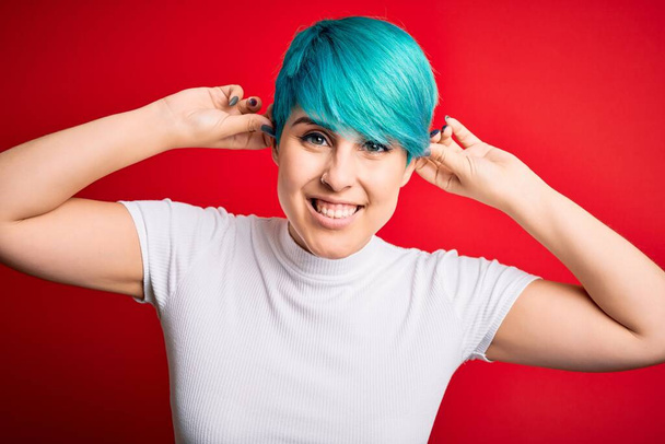 Nuori kaunis nainen sininen muoti hiukset yllään rento t-paita yli punainen tausta Hymyilevä vetämällä korvat sormilla, hauska ele. Koe-esiintyminen - Valokuva, kuva