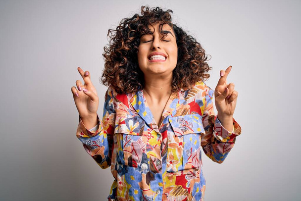 Young beautiful curly arab woman wearing floral colorful shirt standing over white background gesturing finger crossed smiling with hope and eyes closed. Luck and superstitious concept. - Foto, immagini