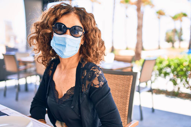 Μέση ηλικία όμορφη μελαχρινή γυναίκα φορώντας γυαλιά ηλίου και ιατρική μάσκα. Συνεδρίαση στο εστιατόριο βεράντα - Φωτογραφία, εικόνα