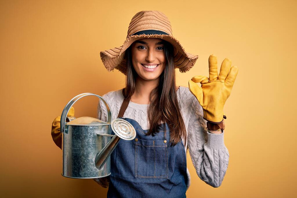 Young beautiful brunette farmer woman wearing apron and hat holding watering can doing ok sign with fingers, excellent symbol - Foto, afbeelding
