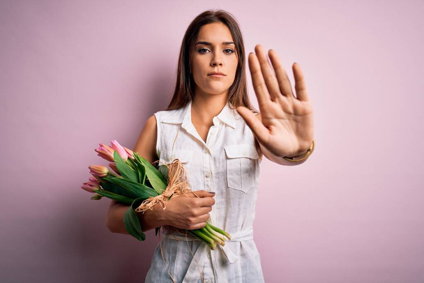 Jonge mooie brunette vrouw met boeket van tulpen bloemen over roze achtergrond doen stoppen met zingen met palm van de hand. Waarschuwingsuitdrukking met negatief en ernstig gebaar op het gezicht. - Foto, afbeelding