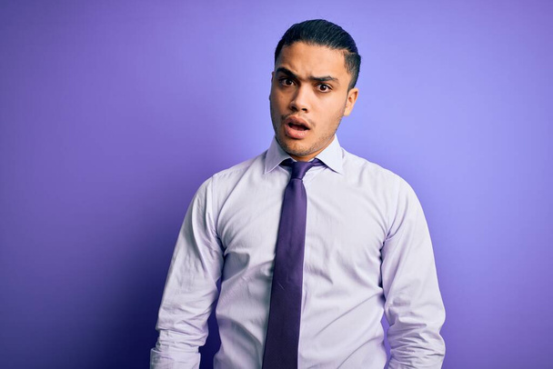 Jonge Braziliaanse zakenman draagt elegante stropdas staande over geïsoleerde paarse achtergrond In shock gezicht, op zoek sceptisch en sarcastisch, verrast met open mond - Foto, afbeelding