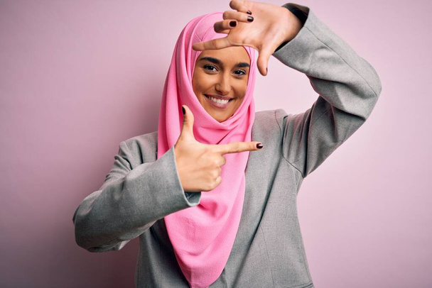 Joven hermosa mujer de negocios morena con hijab musulmán rosa y chaqueta de negocios sonriente haciendo marco con las manos y los dedos con la cara feliz. Concepto de creatividad y fotografía
. - Foto, Imagen