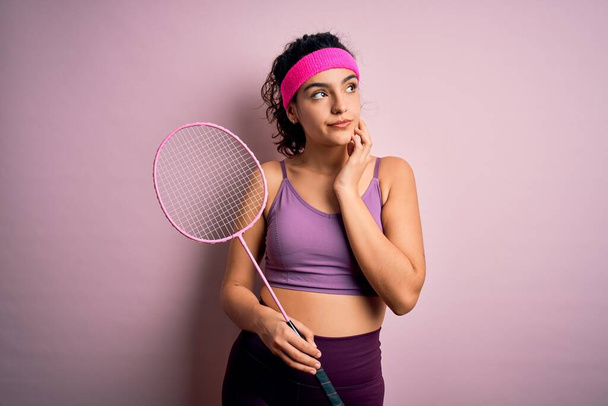 Bonita esportista com cabelo encaracolado vestindo sportswear jogando badminton uning raquete rosto sério pensando em questão, ideia muito confusa
 - Foto, Imagem