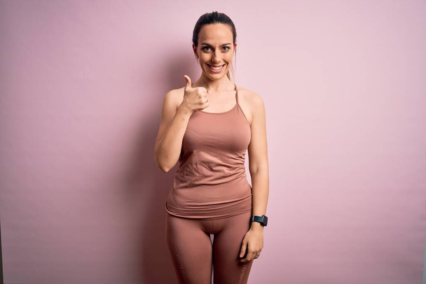 Fiatal szőke fitness nő visel sport edzés ruhák rózsaszín elszigetelt háttér csinál boldog hüvelykujj fel gesztus kézzel. Jóváhagyása kifejezés néz a kamera mutatja siker. - Fotó, kép
