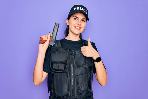Jovem mulher policial vestindo uniforme colete à prova de balas de segurança e segurando arma feliz com grande sorriso fazendo sinal ok, polegar com os dedos, excelente sinal
 - Foto, Imagem