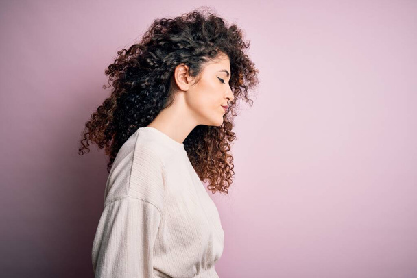 Beautiful woman with curly hair and piercing wearing casual sweater over pink background looking to side, relax profile pose with natural face with confident smile. - Valokuva, kuva
