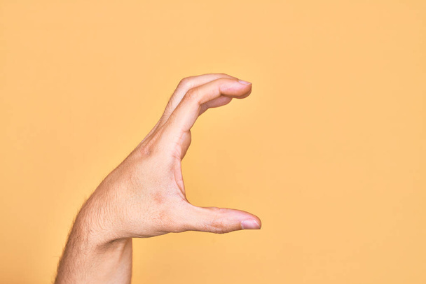 Hand van blanke jongeman met vingers over geïsoleerde gele achtergrond plukken en nemen onzichtbare ding, houden object met vingers tonen ruimte - Foto, afbeelding