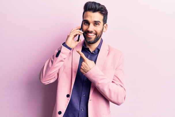 Jeune homme beau avec la barbe avoir la conversation parler sur le smartphone souriant heureux pointant avec la main et le doigt  - Photo, image