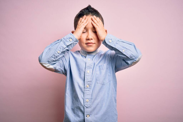 Маленький мальчик в элегантной рубашке, стоящей на розовом изолированном фоне, страдает от головной боли в отчаянии и стрессе из-за боли и мигрени. Руки на голову
. - Фото, изображение