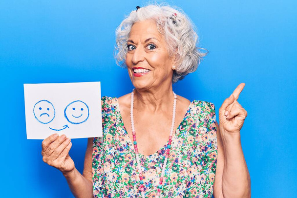 Senior mujer de pelo gris que se entristece por el papel emotivo feliz sonriendo feliz apuntando con la mano y el dedo hacia el lado.  - Foto, Imagen