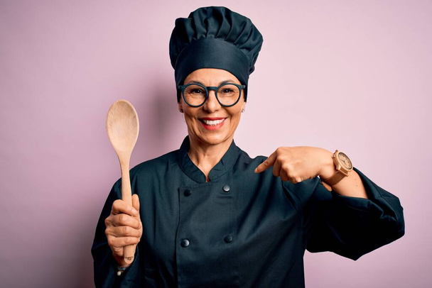 Medioevo bruna chef donna indossa uniforme fornello e cappello tenendo cucchiaio di legno con faccia a sorpresa puntando il dito verso se stesso
 - Foto, immagini
