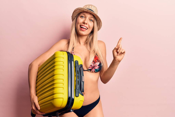 Joven hermosa mujer rubia con bikini y sombrero sosteniendo bolsa de cabina sonriendo feliz señalando con la mano y el dedo a un lado
  - Foto, imagen