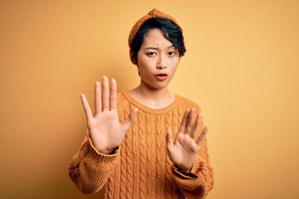 Mladá krásná asijská dívka v ležérním svetru a diadémem stojící nad žlutým pozadím Uhýbající dlaně ukazující odmítnutí a popírání s ustrašeným a nechutným výrazem. Zastavit a zakázat. - Fotografie, Obrázek
