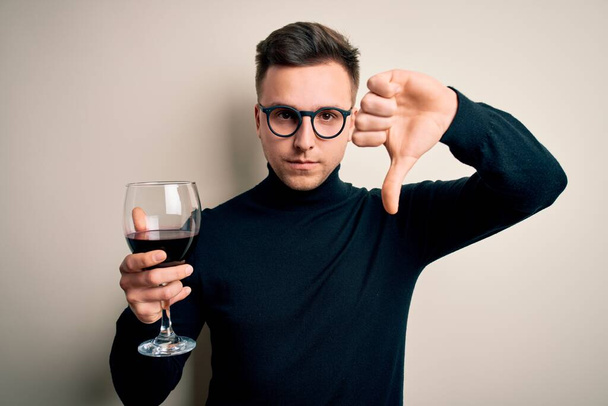 Young handsome caucasian man drinking an alcoholic glass of red wine over isolated background with angry face, negative sign showing dislike with thumbs down, rejection concept - Photo, Image