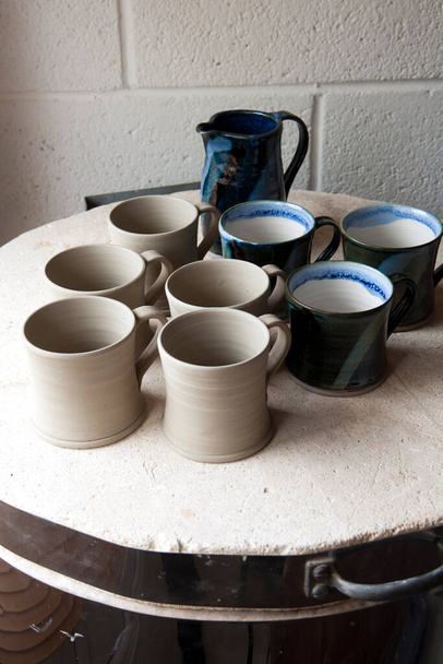 Work in progress standing on the top of the kiln in a ceramics artists home pottery studio. Stoneware mugs before and after. Turned, dried and ready to fire.Glazed inside with white matt glaze, outside dipped with field blue tenmoku. - Photo, Image