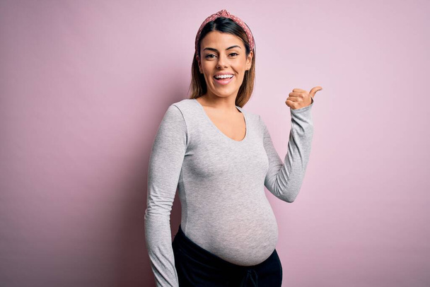 Νεαρή όμορφη μελαχρινή γυναίκα έγκυος περιμένει μωρό πάνω από απομονωμένο ροζ φόντο χαμογελώντας με χαρούμενο πρόσωπο αναζητούν και δείχνοντας προς τα πλάγια με τον αντίχειρα προς τα πάνω. - Φωτογραφία, εικόνα