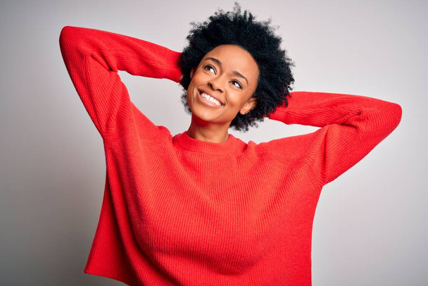 Joven hermosa afroamericana afro mujer con el pelo rizado usando suéter casual rojo relajante y estiramiento, brazos y manos detrás de la cabeza y el cuello sonriendo feliz
 - Foto, Imagen