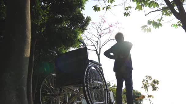 aziatische man oud staan naast rolstoel stretching armen buiten in park - Video