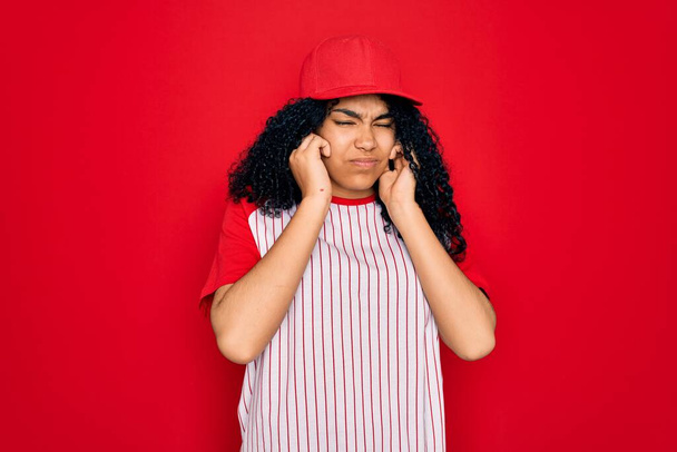 Fiatal afro-amerikai göndör sportos nő baseball sapkában és csíkos póló borító fül ujjakkal bosszantó kifejezés a zaj a hangos zene. Süket elképzelés. - Fotó, kép