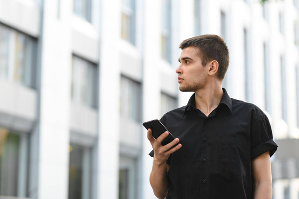 Attractive young office worker in a black shirt stands on a light urban background with a smartphone in his hand and looks away at a blank space. Portrait of stylish office worker - Foto, Imagen