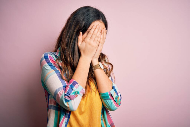 Jonge mooie brunette vrouw dragen casual kleurrijke shirt staan over roze achtergrond met droevige uitdrukking bedekken gezicht met handen tijdens het huilen. Depressieconcept. - Foto, afbeelding