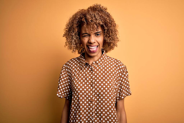 Jeune belle femme afro-américaine portant chemise décontractée debout sur fond jaune clin d'oeil en regardant la caméra avec une expression sexy, gai et heureux visage
. - Photo, image