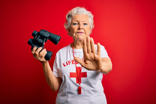 Starszy piękny siwowłosy ratownik kobieta za pomocą lornetki i gwizdać na czerwonym tle z otwartą ręką robi znak stop z poważnym i pewnym siebie wyrazem, gest obrony - Zdjęcie, obraz