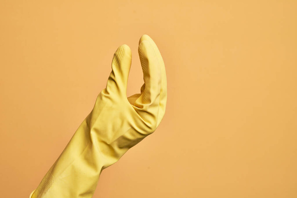 Mano de joven caucásico con guante de limpieza sobre fondo amarillo aislado recogiendo y tomando cosa invisible, sosteniendo el objeto con los dedos mostrando espacio - Foto, imagen