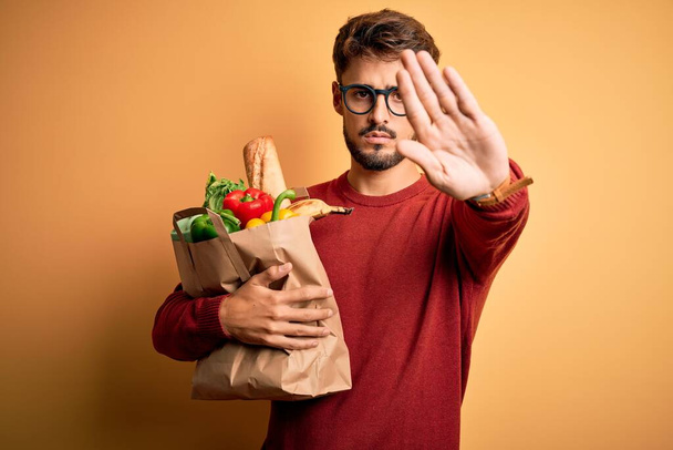 Junger Mann mit Brille hält Papiertüte mit Lebensmitteln über isoliertem gelben Hintergrund mit offener Hand und macht Stoppschild mit ernstem und selbstbewusstem Gesichtsausdruck, Verteidigungsgeste - Foto, Bild