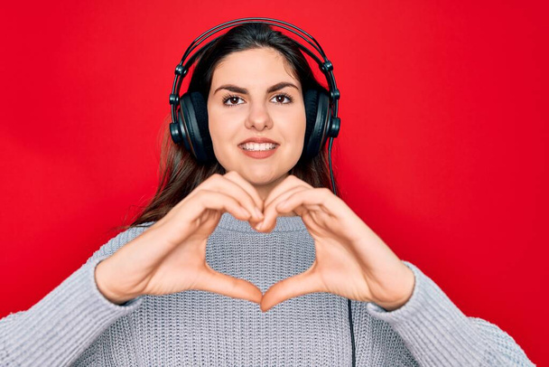 Jong mooi meisje draagt moderne koptelefoon luisteren naar muziek over rode achtergrond glimlachen in liefde tonen hart symbool en vorm met de handen. Romantisch concept. - Foto, afbeelding