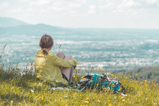 Sporty girl on a hiking trip is sitting on the meadow an enjoying the view over the far away city - Foto, Imagen