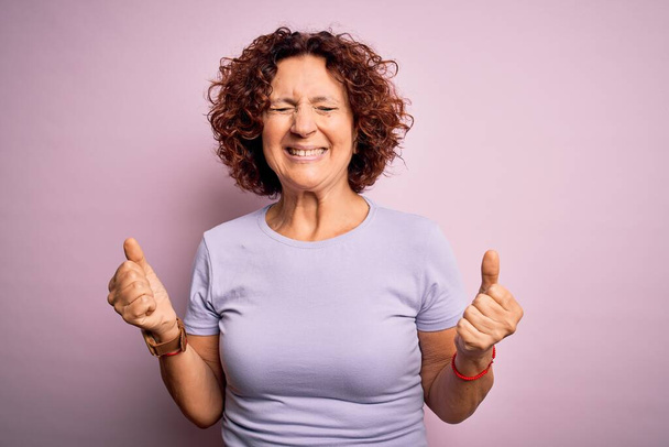 Schöne Frau mittleren Alters mit lockigem Haar trägt lässiges T-Shirt über isoliertem rosa Hintergrund, sehr glücklich und aufgeregt dabei Siegergeste mit erhobenen Armen, lächelnd und schreiend nach Erfolg. Festkonzept. - Foto, Bild
