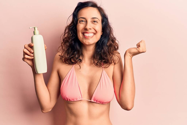 Young beautiful hispanic woman wearing bikini holding sunscreen lotion screaming proud, celebrating victory and success very excited with raised arm  - Фото, зображення