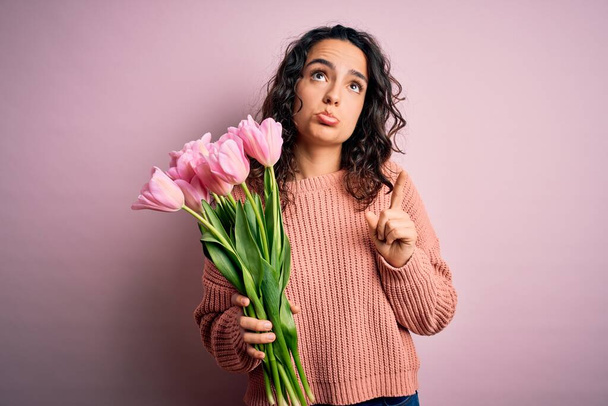 Mladá krásná romantická žena s kudrnatými vlasy, držící kytici růžových tulipánů, ukazující smutně a rozrušeně, ukazující na směr prsty, nešťastná a sklíčená. - Fotografie, Obrázek