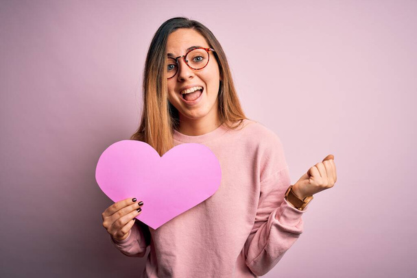 Young beautiful blonde romantic woman wearing glasses holding big pink heart screaming proud and celebrating victory and success very excited, cheering emotion - Photo, Image