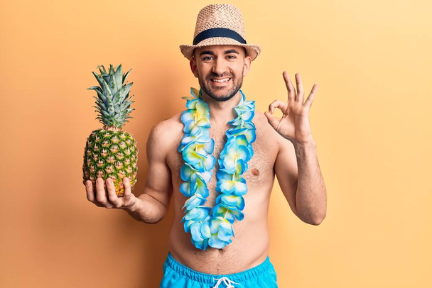 Jonge knappe kale man draagt zwemkleding en Hawaïaanse lei houdt ananas vast doet het goed teken met vingers, glimlachend vriendelijk gebaar uitstekend symbool  - Foto, afbeelding