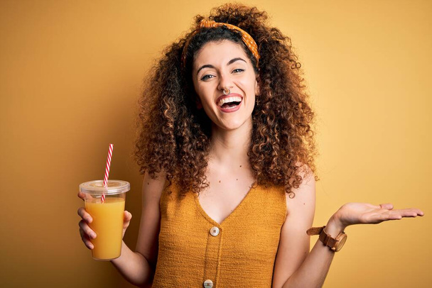 Молодая красивая женщина с вьющимися волосами и пирсинг пить здоровый апельсиновый сок очень счастливый и взволнованный, победитель выражение празднования победы кричать с большой улыбкой и поднятые руки
 - Фото, изображение