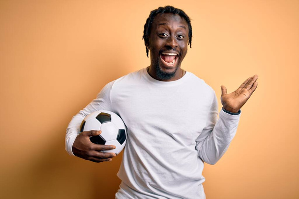 Giovane giocatore afro-americano uomo che gioca a calcio tenendo palla da calcio su sfondo giallo molto felice ed eccitato, espressione vincitore celebrando la vittoria urlando con grande sorriso e mani alzate
 - Foto, immagini