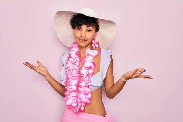Joven hermosa mujer turista afroamericana con bikini y flores de lei hawaiana expresión despistada y confusa con los brazos y las manos levantadas. Concepto de duda
. - Foto, imagen