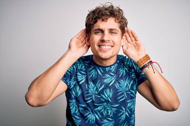 Young blond handsome man on vacation with curly hair wearing casual summer t-shirt Trying to hear both hands on ear gesture, curious for gossip. Hearing problem, deaf - Photo, Image