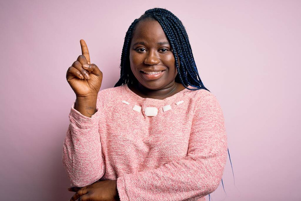 afroamericano plus size donna con trecce indossa maglione casual su sfondo rosa mostrando e indicando con dito numero uno, mentre sorridente fiducioso e felice
. - Foto, immagini