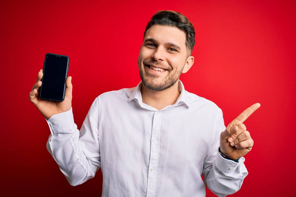 Giovane uomo d'affari con gli occhi blu che mostra lo schermo dello smartphone su sfondo rosso molto felice che punta con mano e dito di lato
 - Foto, immagini