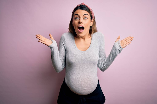 Young beautiful brunette woman pregnant expecting baby over isolated pink background celebrating crazy and amazed for success with arms raised and open eyes screaming excited. Winner concept - Photo, Image