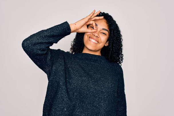 Junge afrikanisch-amerikanische lockige Frau in lässigem Pullover, der über weißem Hintergrund steht und ok Geste mit lächelnden Händen macht, Augen, die durch Finger mit glücklichem Gesicht schauen. - Foto, Bild