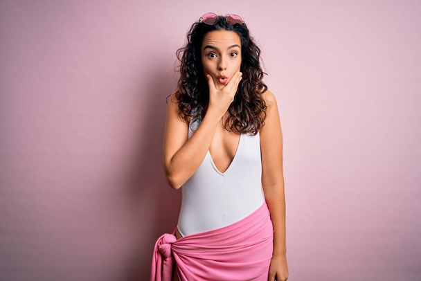 Beautiful woman with curly hair on vacation wearing white swimsuit over pink background Looking fascinated with disbelief, surprise and amazed expression with hands on chin - Photo, Image